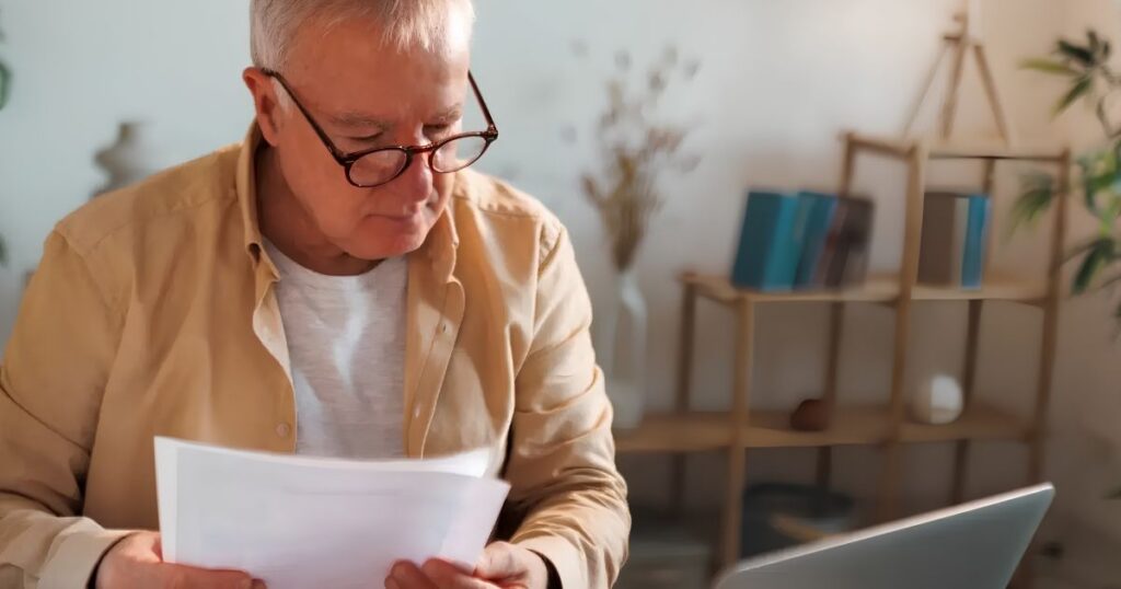 Older man with glasses reading documents at a desk in a cozy home office setting.