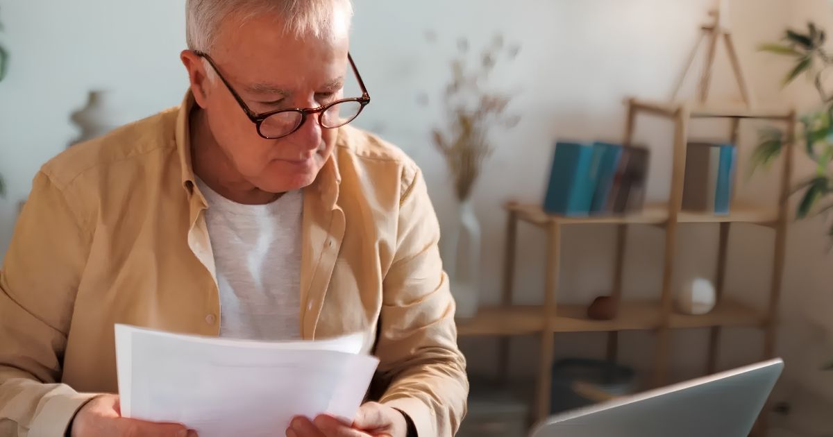 Older man with glasses reading documents at a desk in a cozy home office setting.
