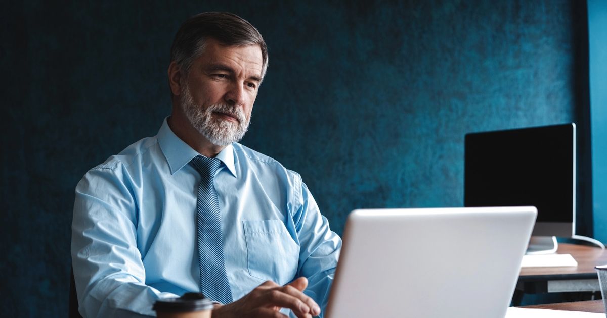 Older man with gray beard in a blue shirt and tie sits at a desk, using a laptop in a blue-walled office.