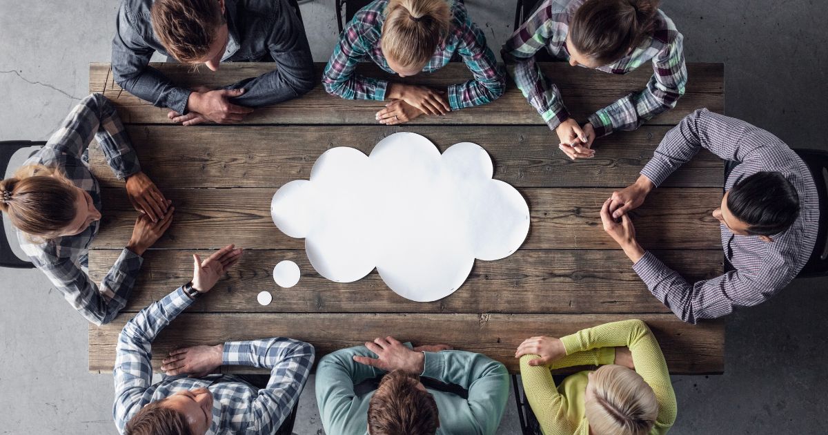 Eight people sit around a wooden table in a circle, a large cloud thought bubble sits in the center, signaling brainstorming.