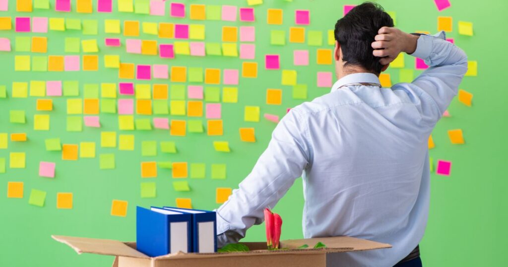 Man stands with back to camera in a cardboard box, scratching his head as colorful sticky notes cover a green wall, suggesting brainstorming or planning.