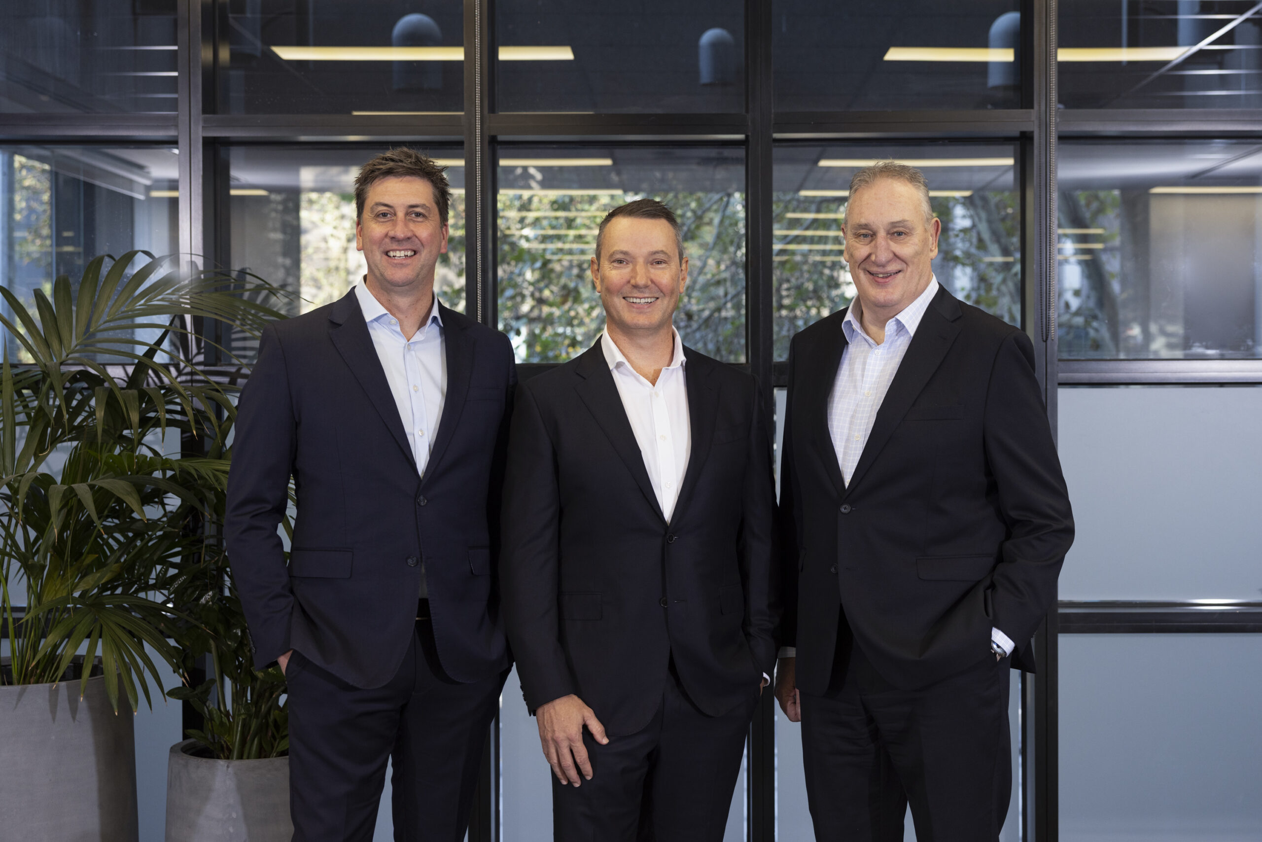 Three men in dark suits posing and smiling in a modern office lobby with glass partitions and plants behind them.