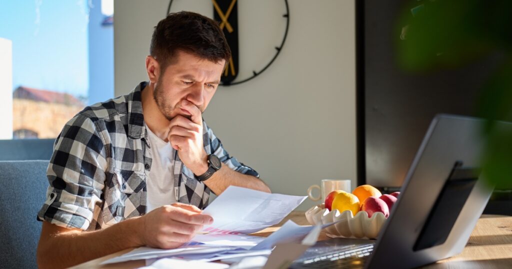 Man in a plaid shirt studies documents at a home office, with a laptop and a fruit bowl on the desk.