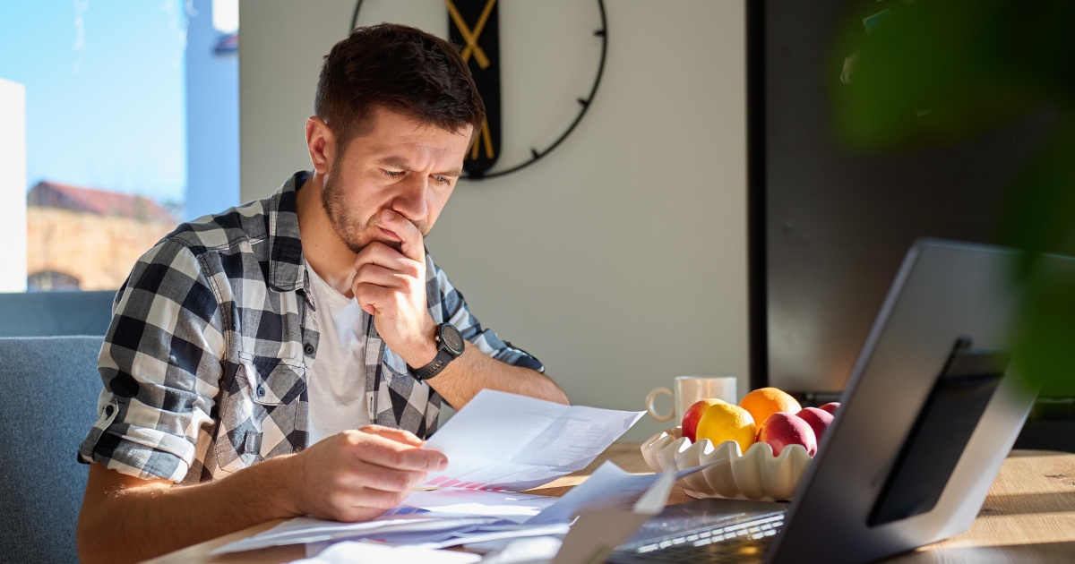 Man in a plaid shirt studies documents at a home office, with a laptop and a fruit bowl on the desk.