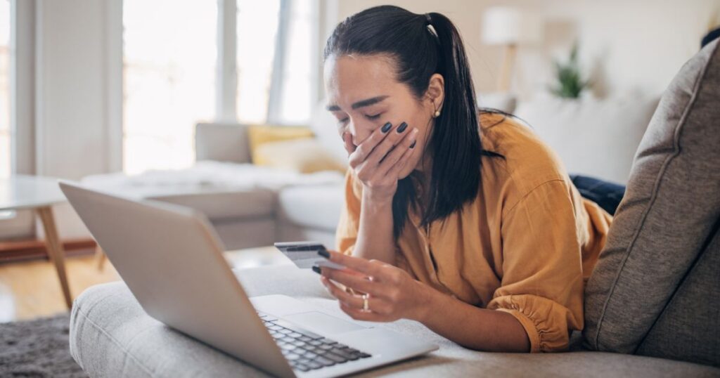 Woman lounging on a sofa, holding a credit card while looking at a laptop, appearing anxious about online payment.