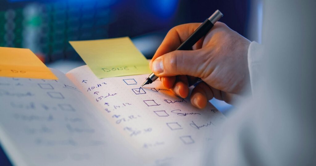 Close-up of a hand writing in a dotted notebook, checking off items on a checklist with sticky notes nearby