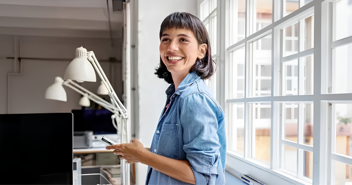 Smiling woman in a denim shirt stands by a bright office window, holding a smartphone.