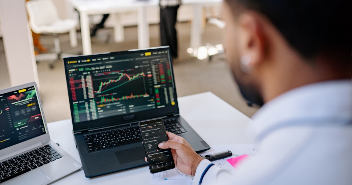 A trader monitors stock charts on two laptops while checking a trading app on a smartphone at a bright desk.