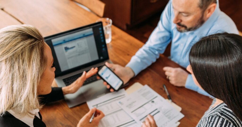 Three professionals review documents at a wooden table, with a laptop and smartphone in use.
