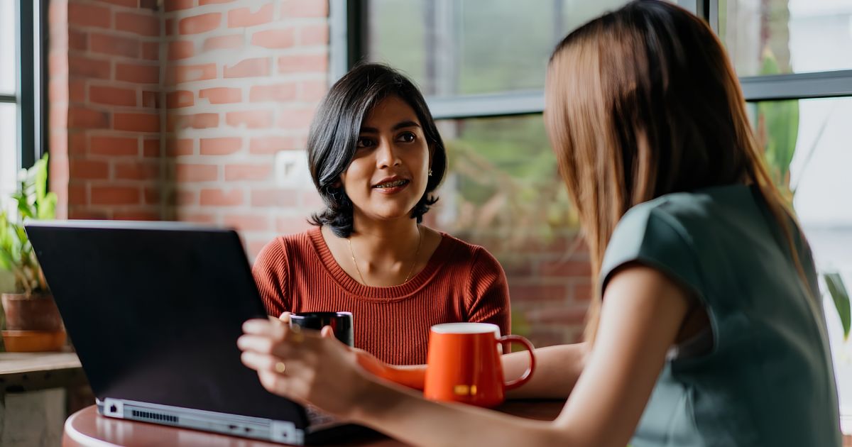 Two women converse at a cafe table with an open laptop and orange mugs nearby.