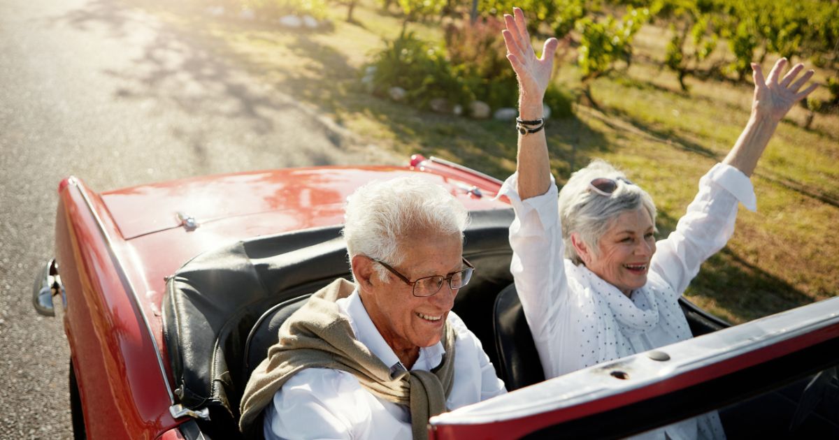 Couple in a red convertible car smiling, woman raising arms in celebration on a sunny countryside road.