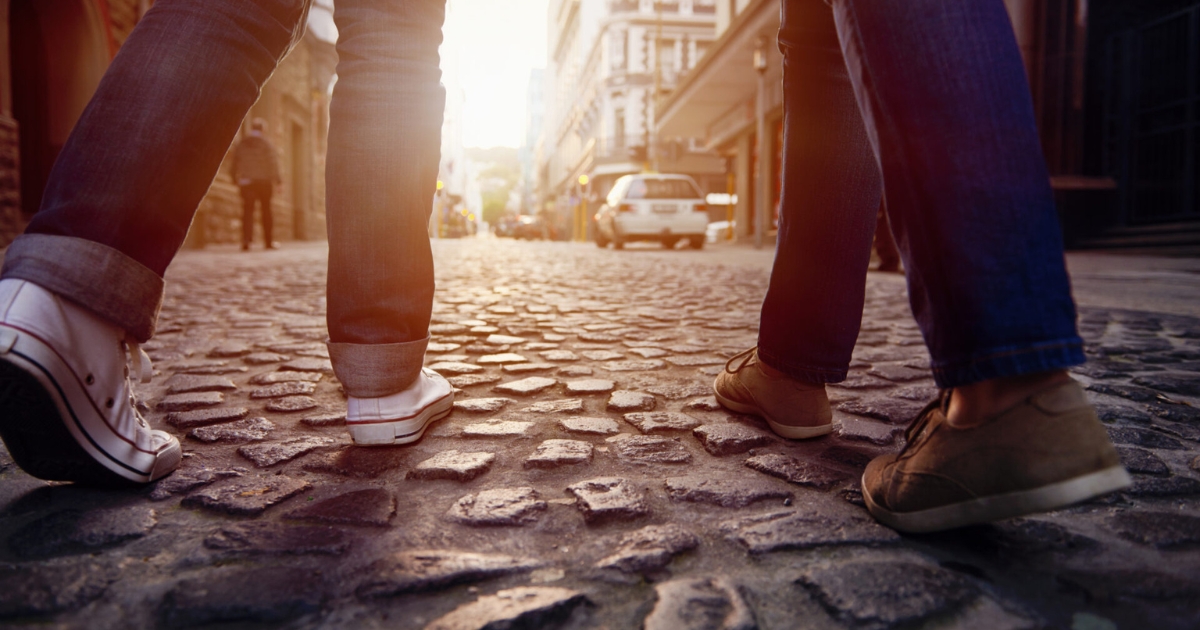 Feet in sneakers and casual shoes stepping on uneven cobblestones in a city street at sunset, viewed from ground level.